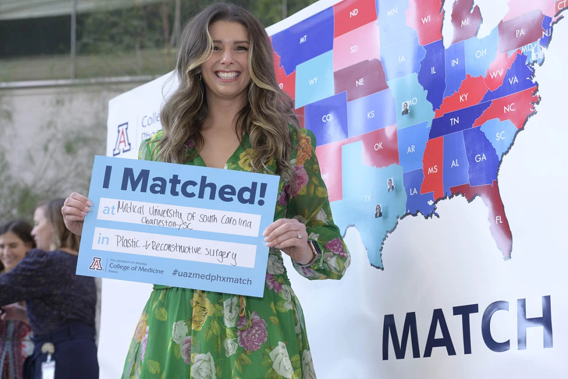 Young woman in dress and long brown hair holds a poster that says "I matched" in front of a map of the U.S.