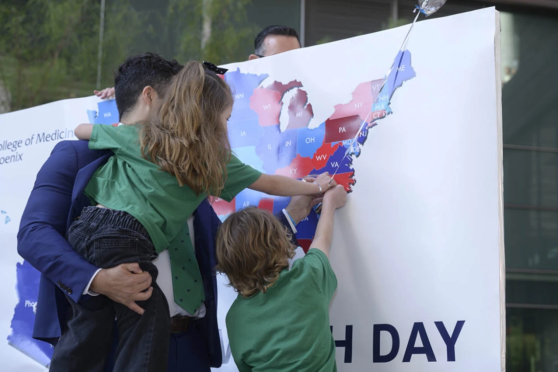 A man in a blue suit holding a girl and next to a boy stick a pin in a map of the U.S.