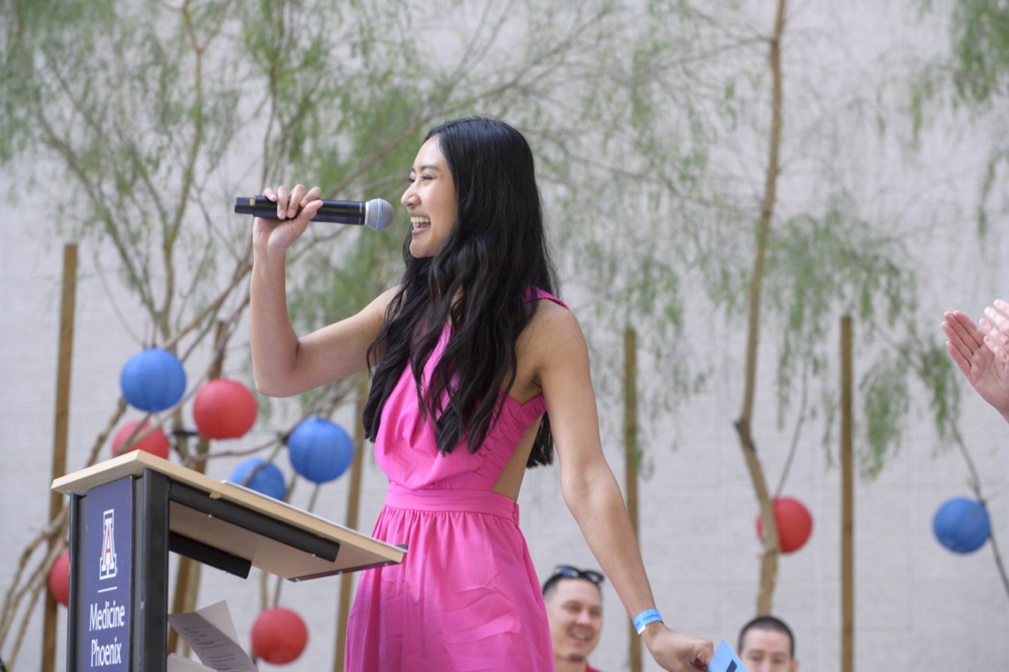 A young Asian woman with long dark hair speaks into a microphone in an outdoor setting. 