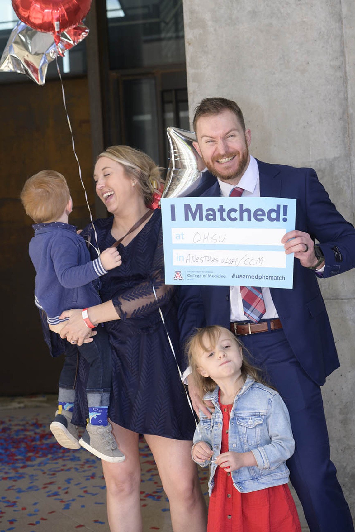 A family stands together as the dad holds an "I Matched" sign and his wife looks at the boy she is holding and smiles. Young girl standing in front of them.