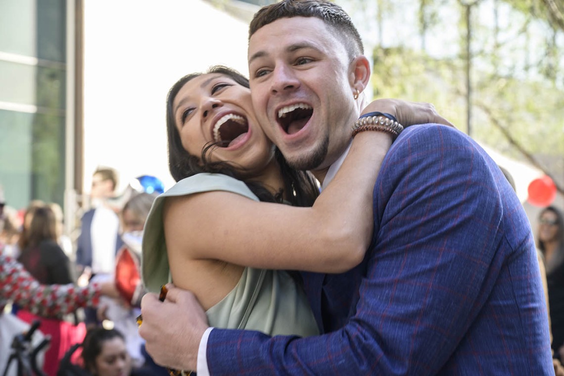 A young woman and man with light skin hug each other with huge open-mouthed smiles.