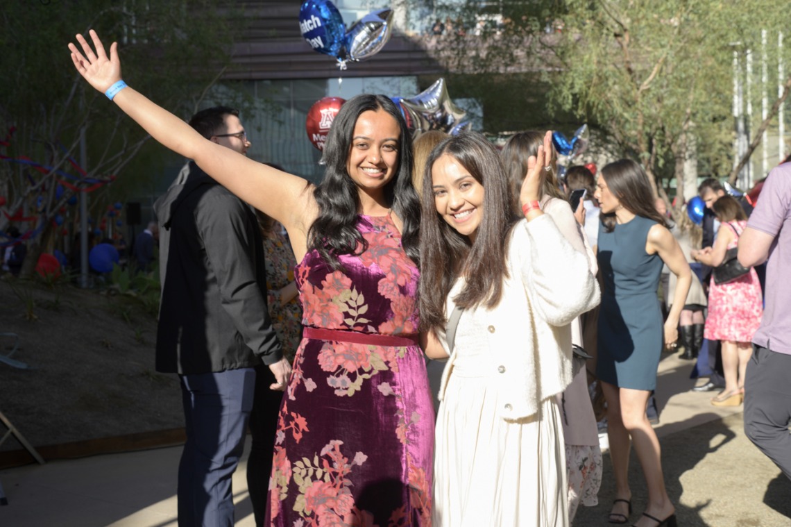 Two young woman stand side by side smiling and waiving. Both have long dark hair and light brown skin. 