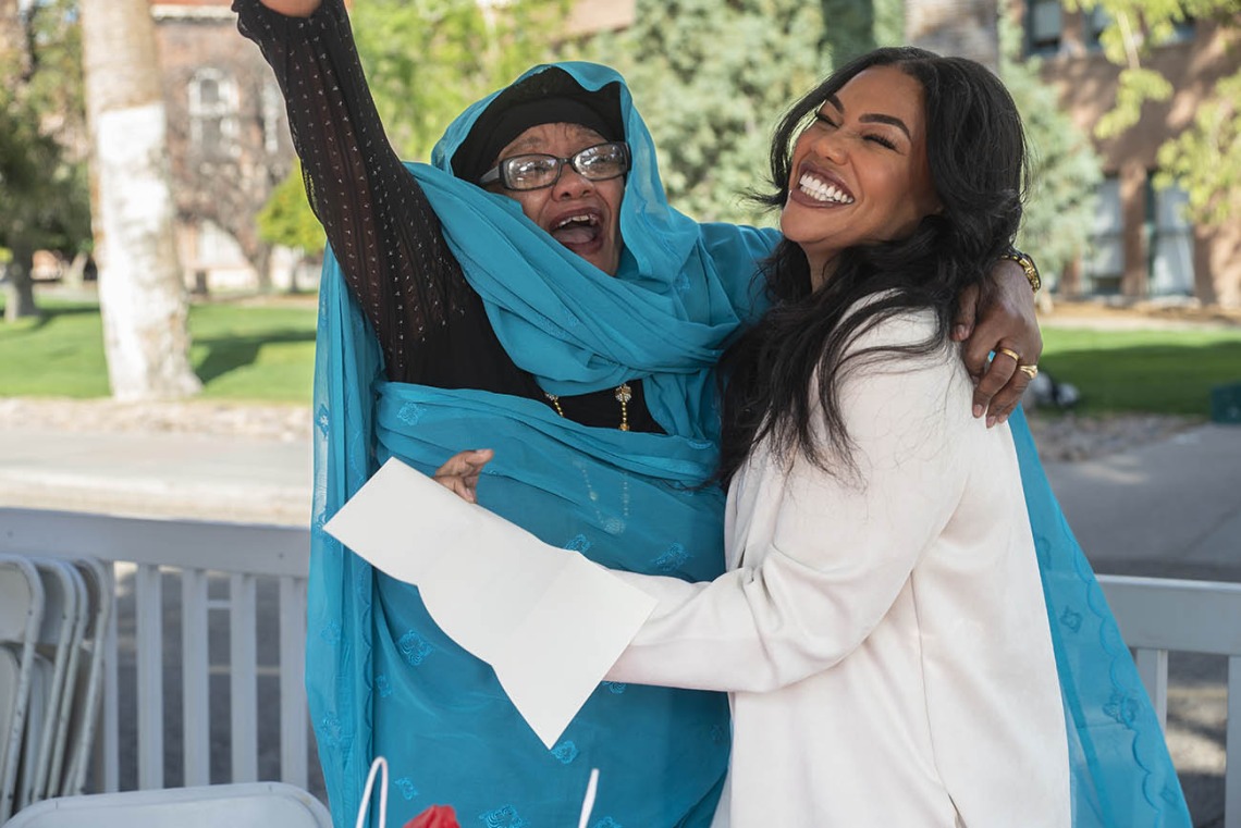 An older dark-skinned woman smiles and holds up one arm in celebration as she hugs a smiling younger dark-skinned woman. 