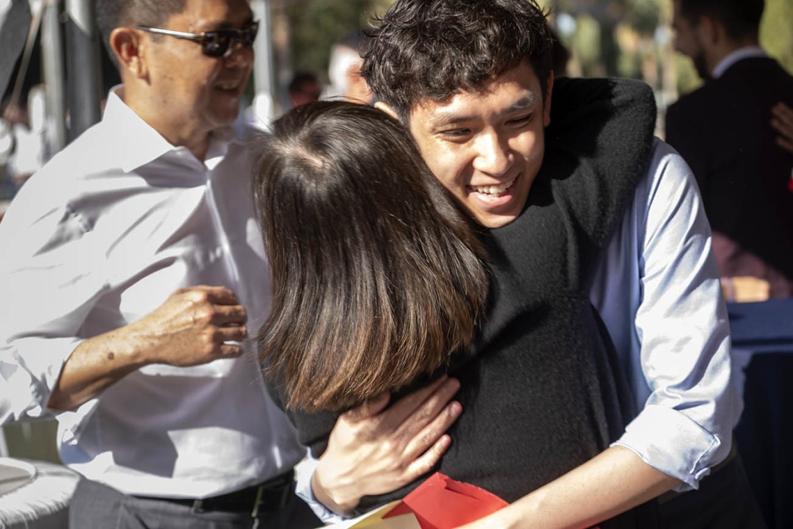 A young man with wiht a big smile and curly dark hair hugs his mom.