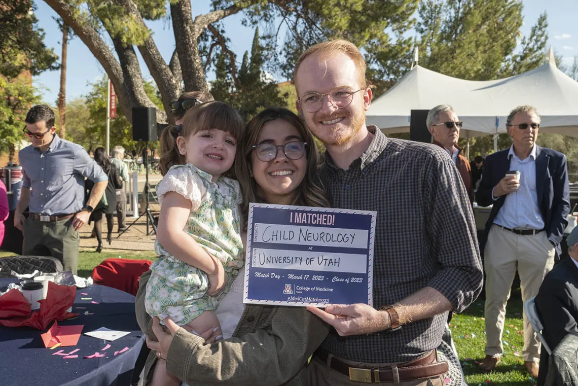 A young couple holding a little girl smile while holding a Match Day sign. All three are light skinned.