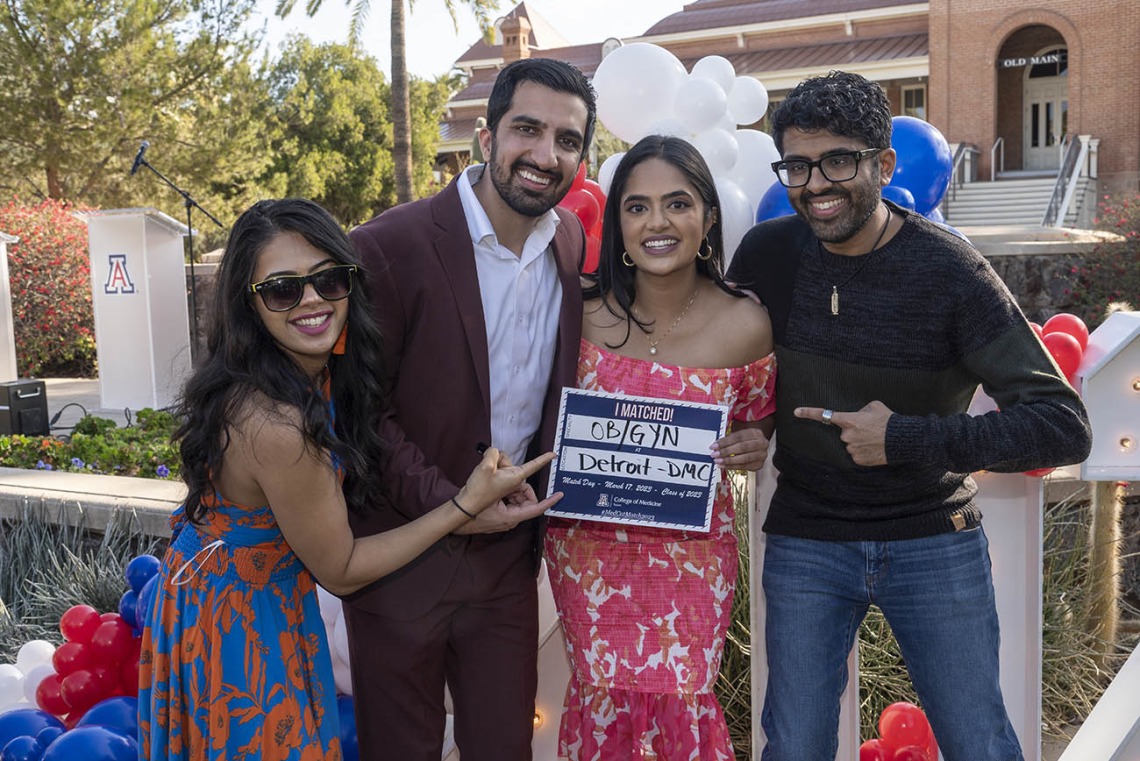 Four young adults, two men and two women, with dark skin and hair smile while the woman in the center holds a sign signifying where she matched.
