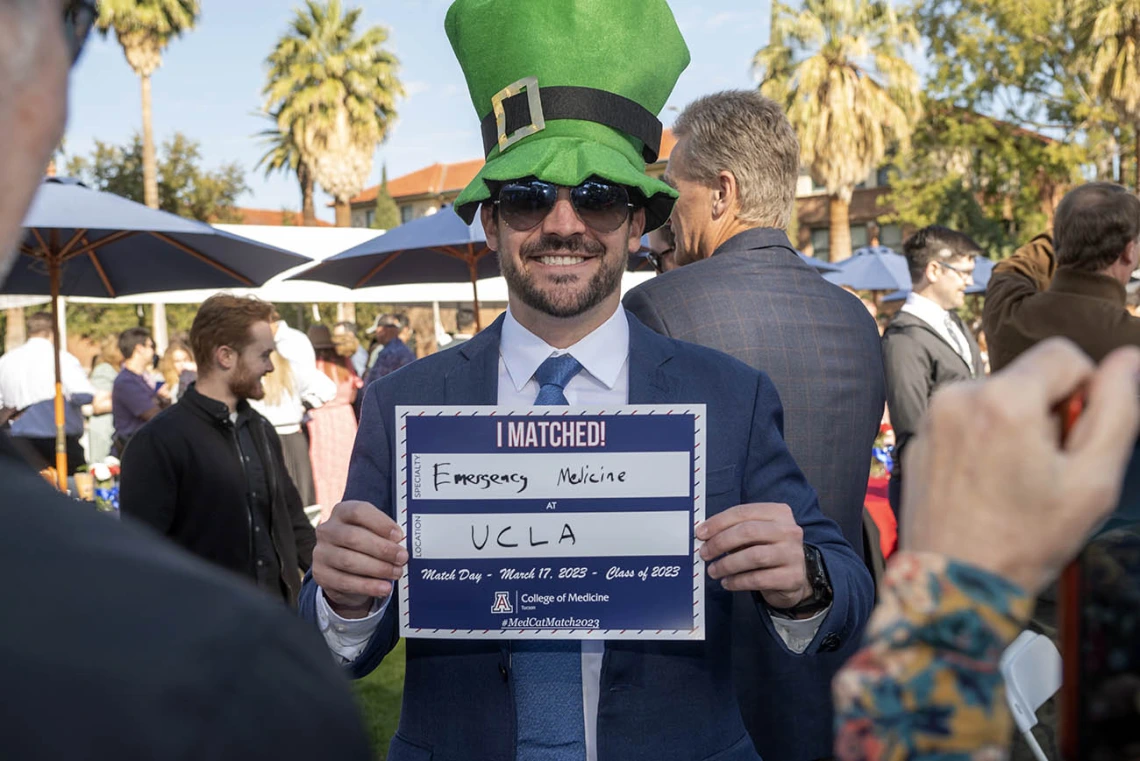 A man with a beard wearing a suite and big green hat hods a sign that says "I matched at UCLA"