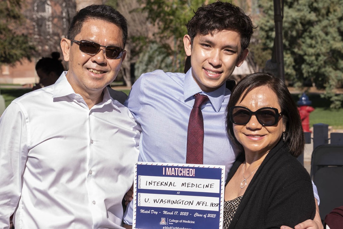 A young Asian man with a dress shirt and tie poses between his mom and dad. They are holding up a sign saying where he matched. 