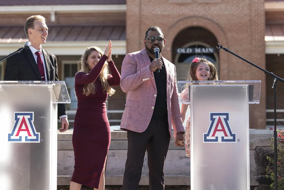 A man in a light pink jacket speaks into a microphone while holding the hand of a young girls who is smiling. 