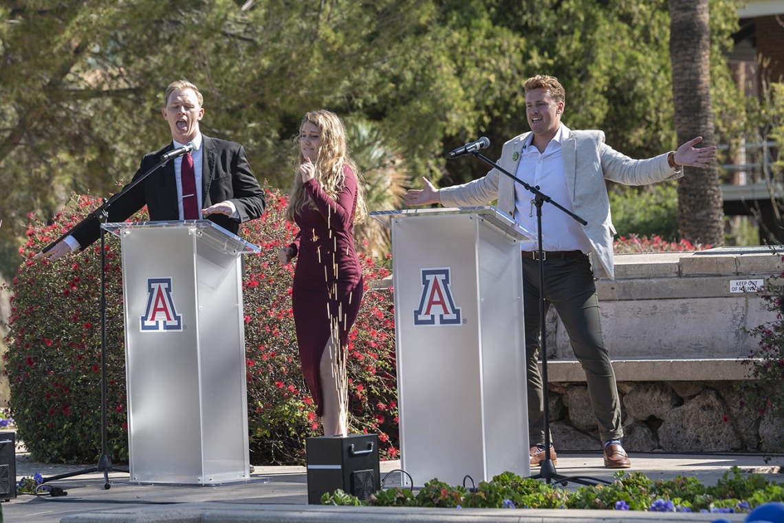 Two men and a woman stand on a stage with two podiums with UArizona logo. Man on right has his hands outstreached and is smiling. 