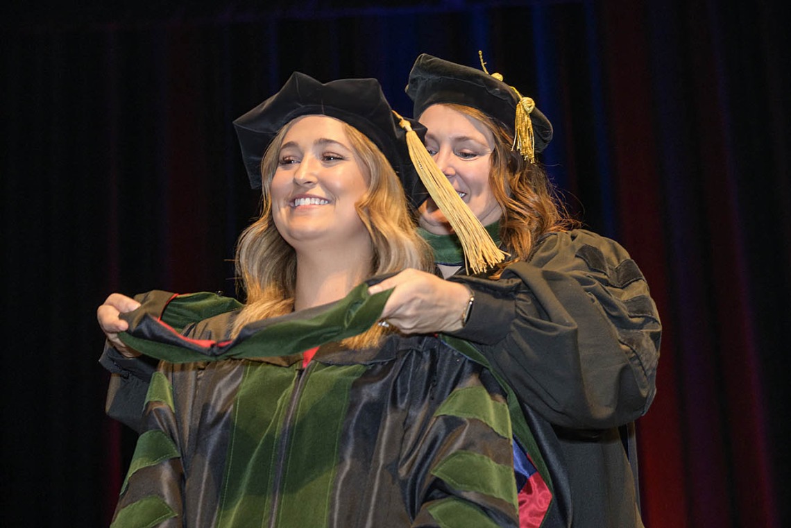 A young blonde woman in graduation regalia smiles as a professor places a sash over her shoulders. 
