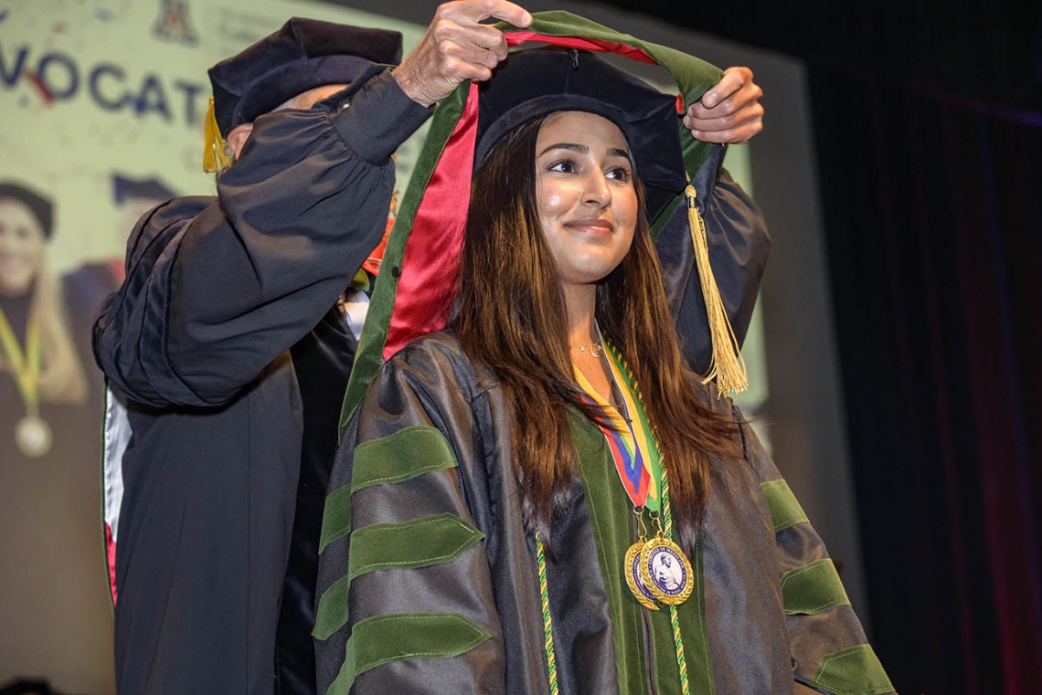 A young woman with long dark hair wearing a cap and gown has a sash placed over her shoulders. 