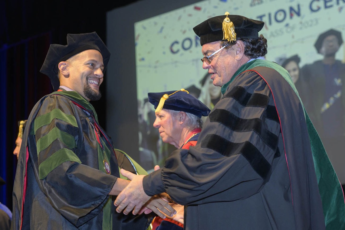 A smiling young man in graduation regalia shakes hands with the dean of the college. 