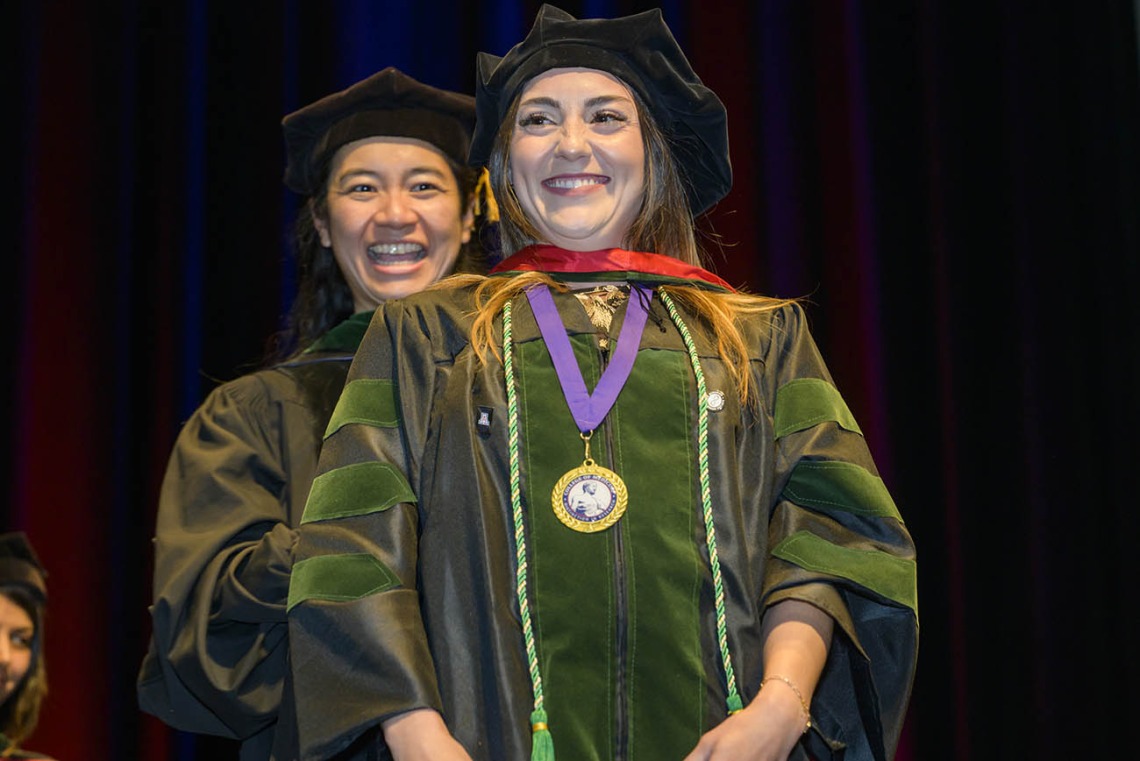 A young woman with long dark hair wearing a graduation cap and gown smiles as a faculty member behind her smiles as well. 
