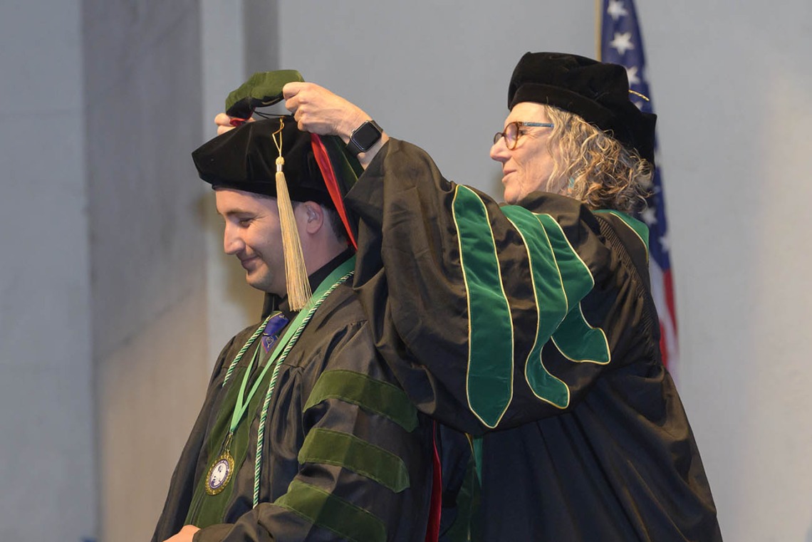Side view of a young man in graduation regalia has a sash placed over his head by a female professor, alos in graduation regalia.