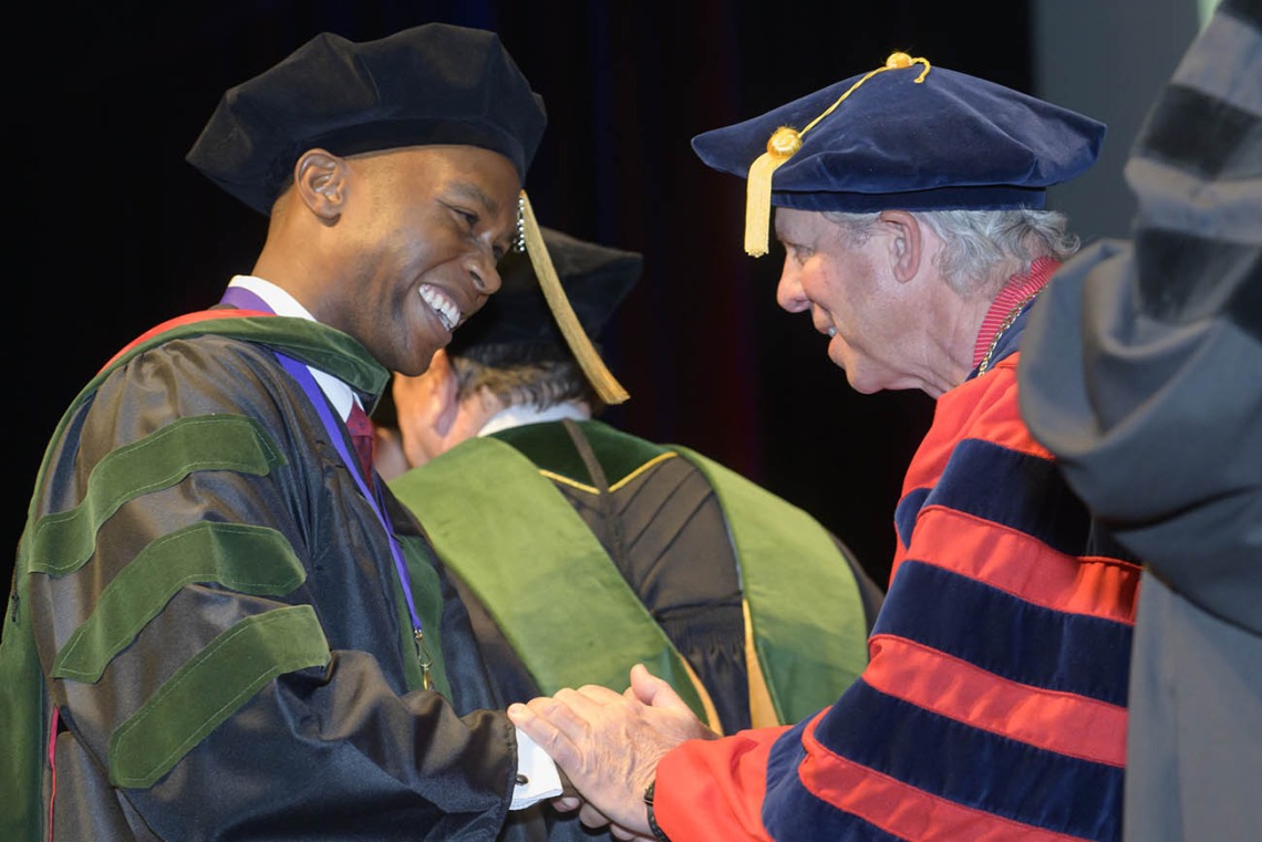 A young man in graduation cap and gown smiles as he shakes hands with the president of the university, also in graduation regalia.