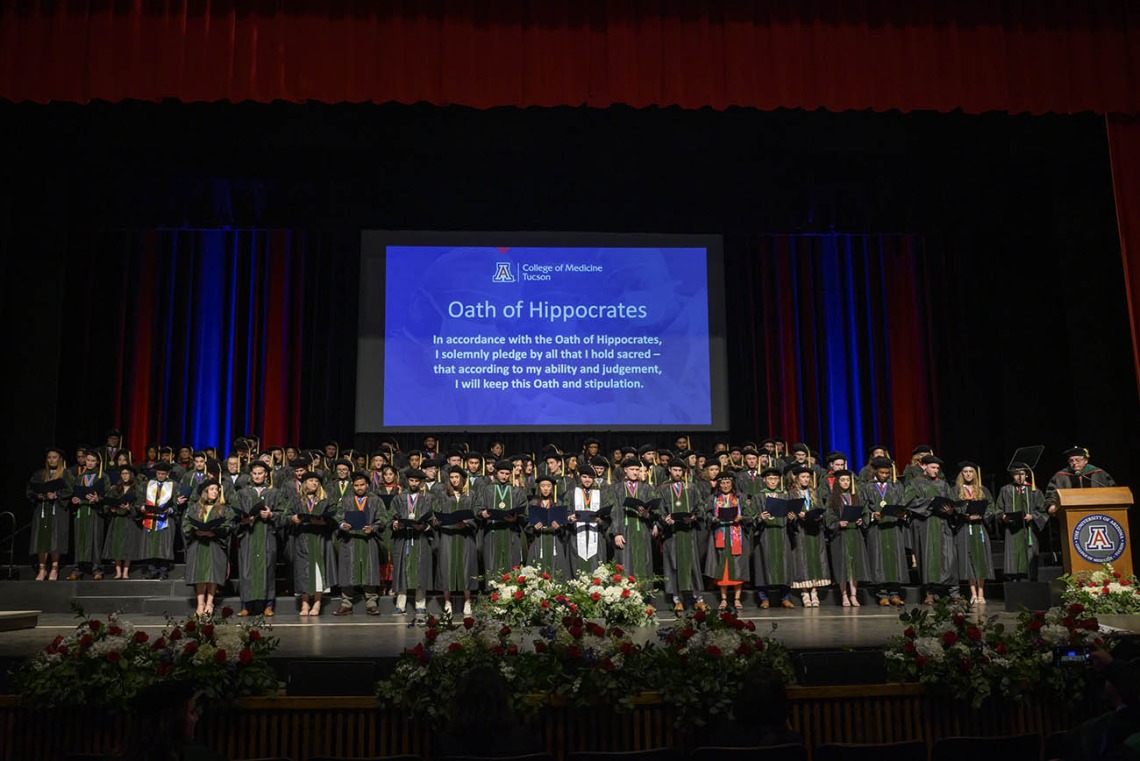 Over 100 medical school graduates in caps and gowns stand on a stage under a large blue display screen with an oath on it. 