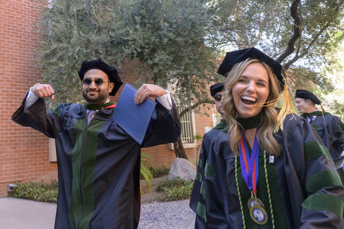 A young man and woman in graduation regalia smile and gesture in celebration as they walk outside. 