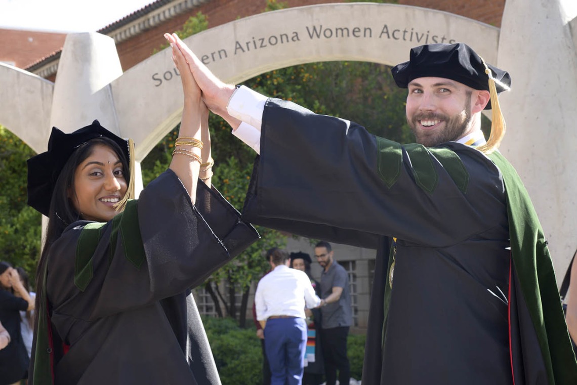 A young woman and man in graduation robes and hats smile as they hold their hands together overhead to make an arch. 