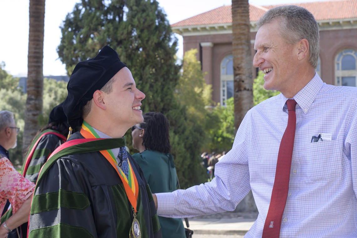 A young male graduate wearing a cap and gown faces an older man who is patting him on the arm in congratulations. 