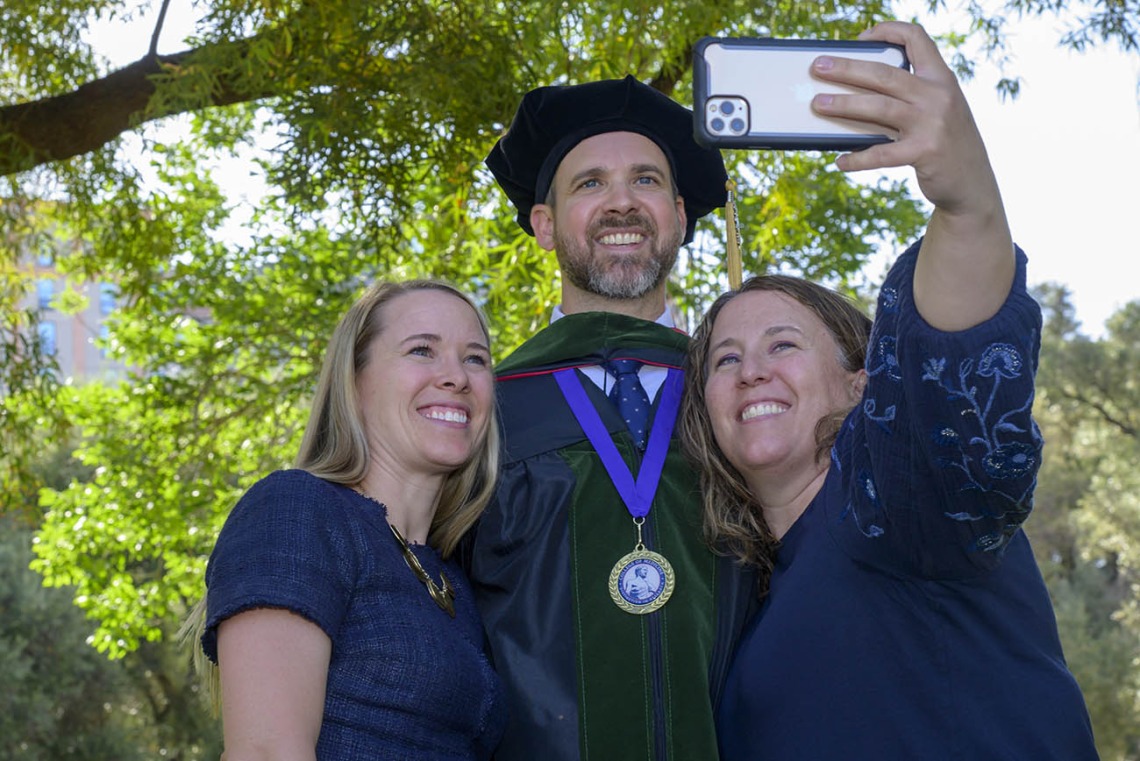 Two women stand on each side of a tall man in graduation cap and gown as one of the women takes a selfie. 