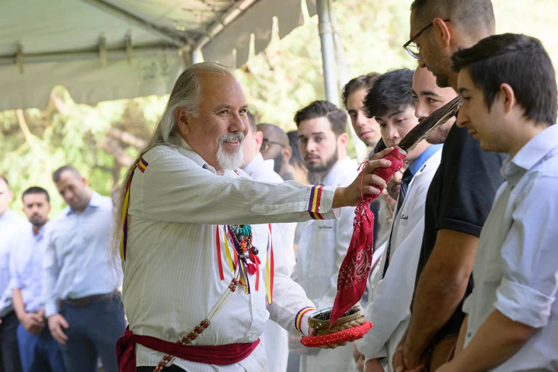 An older man in a traditional Native American designed shirt touches the forehead of a young man with an eagle feather. 