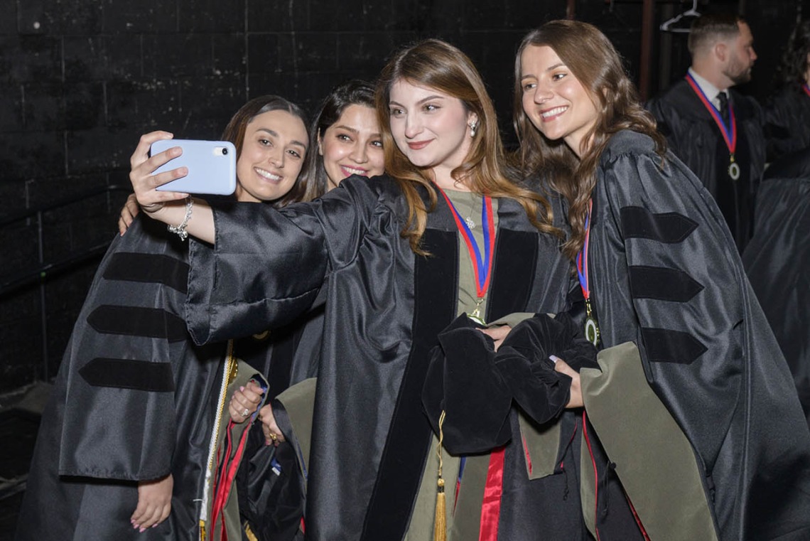 Four young women in black graduation caps and gowns smile as they take a selfie.