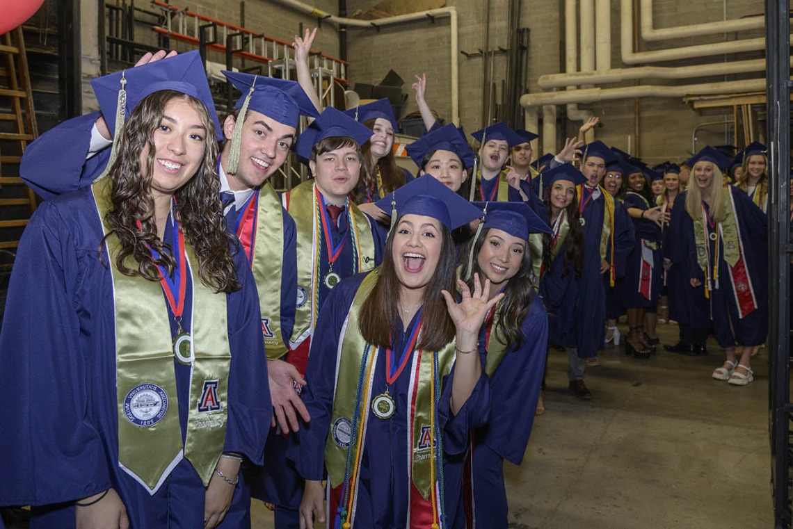 A large group of graduates in blue caps and gowns smile and cheer in a line backstage before their graduation ceremony. 