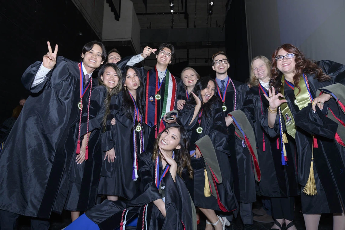 About a dozen pharmacy graduates in caps and gowns bunch together smiling and cheering.