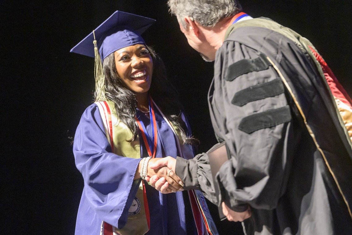 A young Black woman in a blue graduation cap and gown smiles as she shakes hands with a professor. 