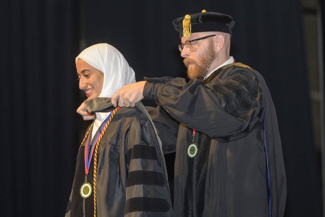 A young woman wearing a white head scarf and a graduation gown has a sash placed over her shoulders by professor.