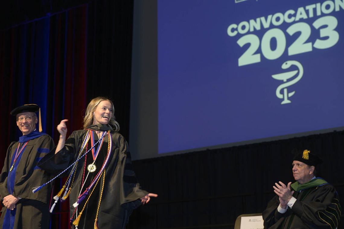 A young white woman in graduation gown bounds across the stage in front of a large screen that reads "Convocation 2023"
