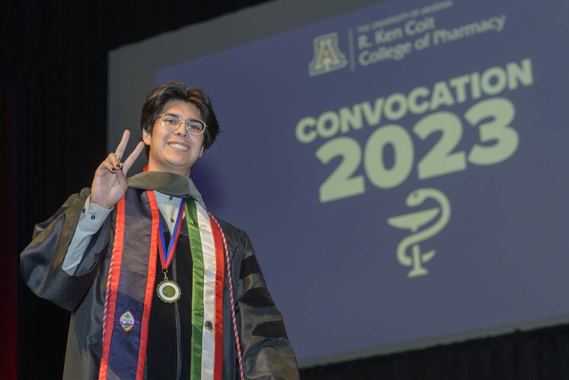 A young man in a graduation gown flashes a peace sign and smiles as he crosses the stage. 