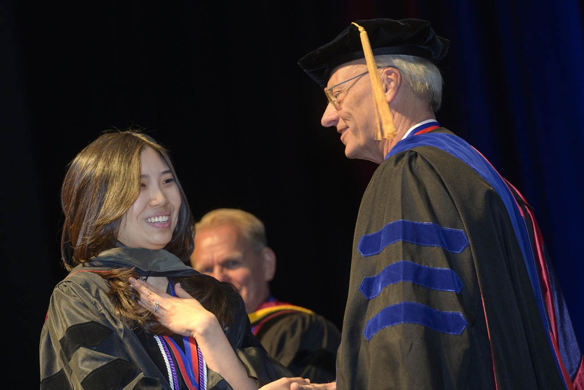 A young Asian woman in a graduation gown shakes hands with an older white man, also in cap and gown. 