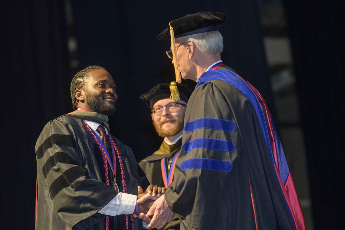 A young Black man in a graduation gown shakes hands with the college dean, an older white man.