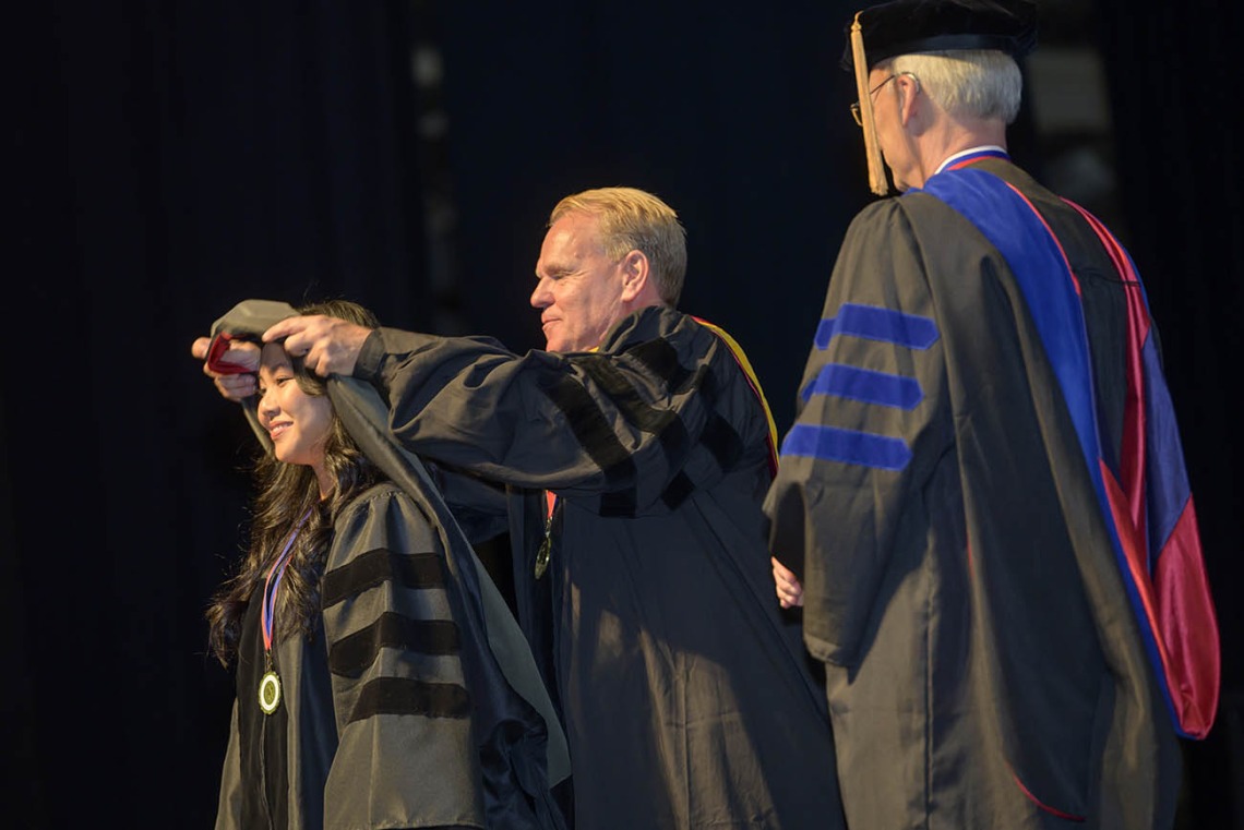 A young Asian woman in a graduation gown has a sash placed over her shoulders by a middle-aged white professor in a black graduation gown. 
