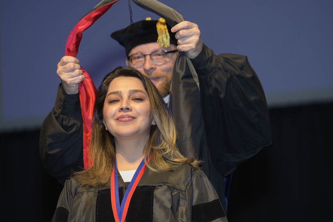 A young woman with light brown hair wearning a graduation gown smiles as a professor places a sash over her shoulders. 