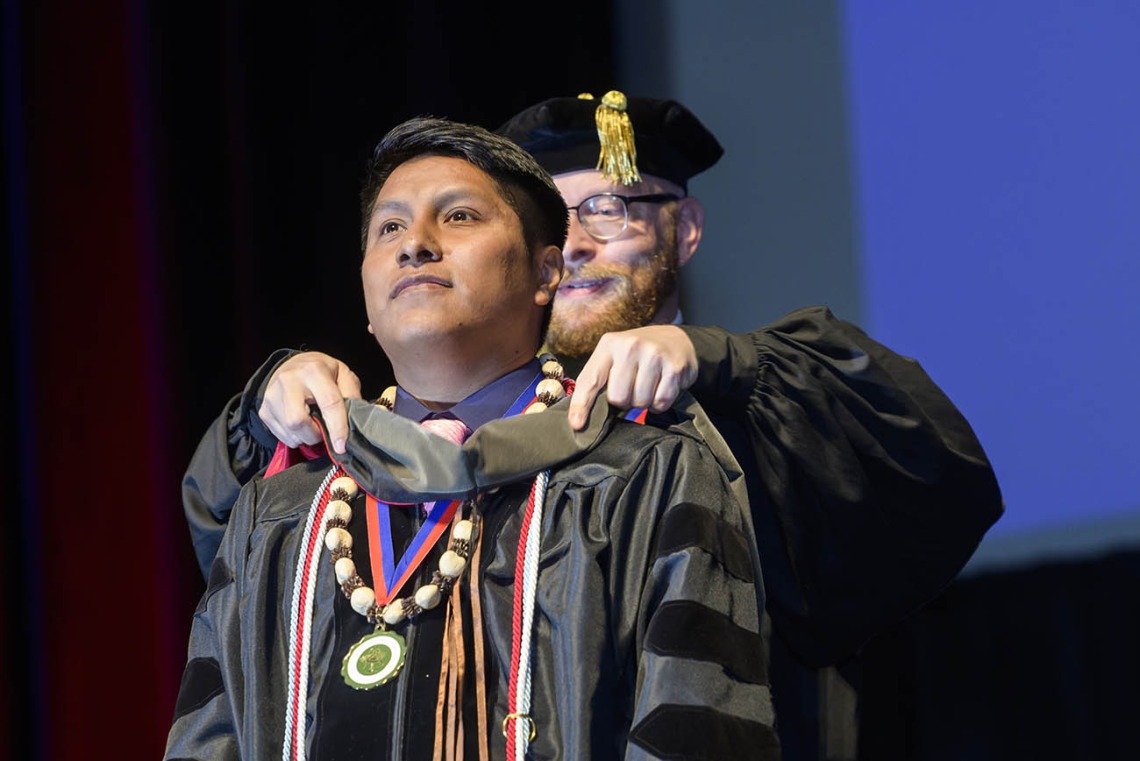 A young Native American man in a graduation gown and beaded necklace has a sash placed over his shoulders by a professor. 