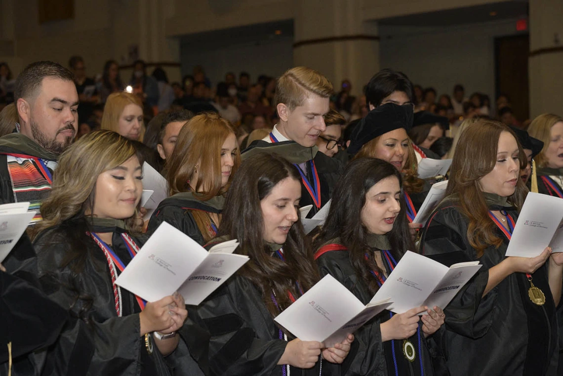 A large group of Pharmacy graduates in graduation gowns read from pamphlets they are all holding. 