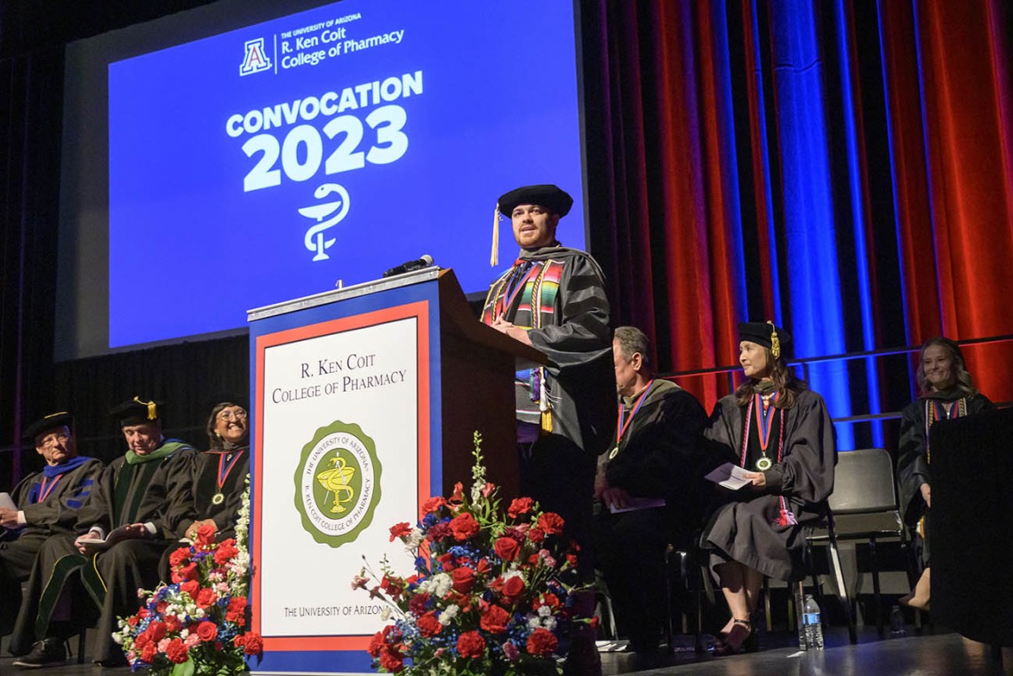 A young man in a black graduation cap and gown stands on a stage at a podium with a screen behind him that says "Convocation 2023"