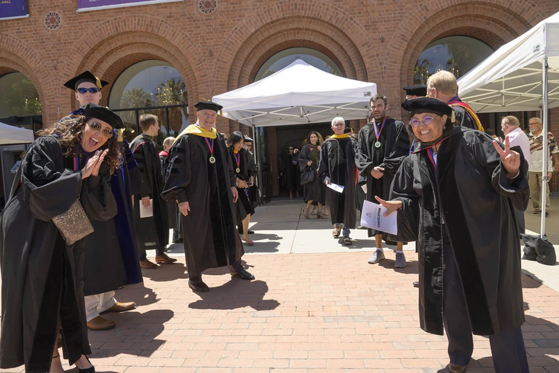 A group of Pharmacy professors stand on both sides of a walkway outside a large brick building waving and cheering. 