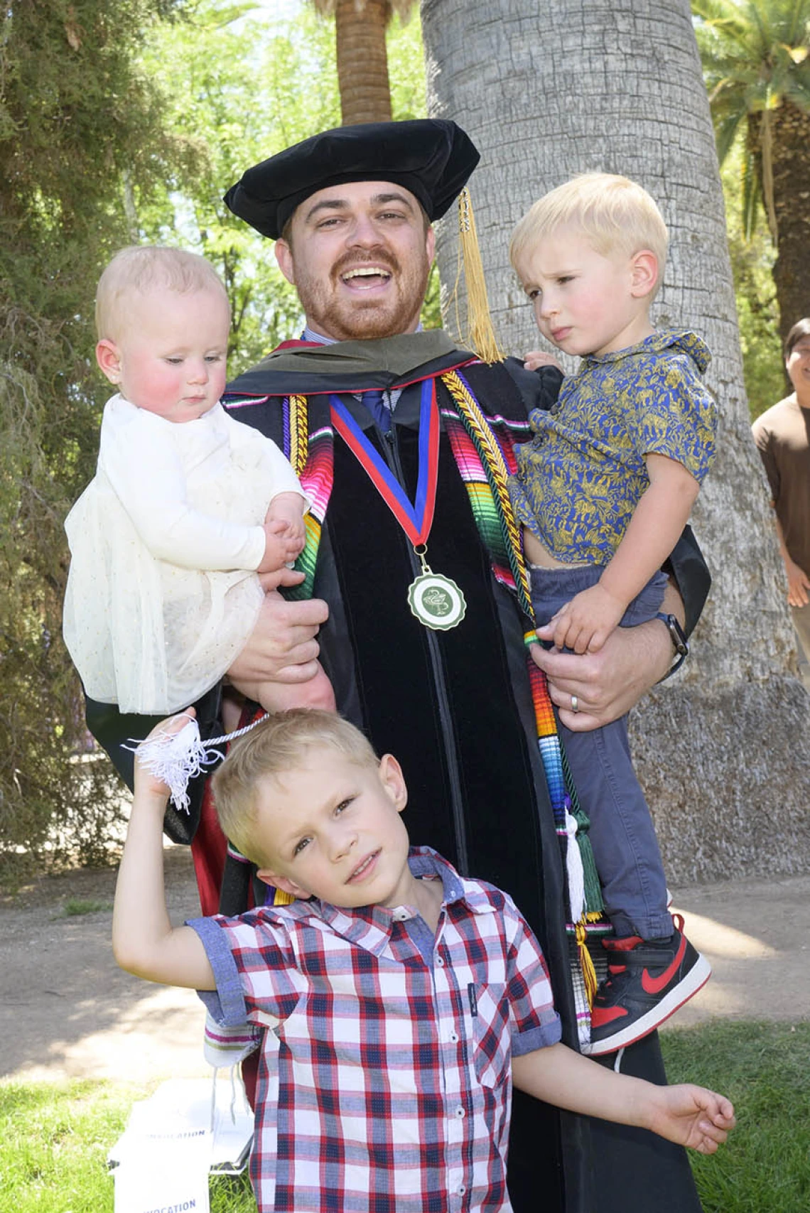 A young man with a beard wearing a graduation cap and gown holds two young children in his arms while another child stands in front of him. 