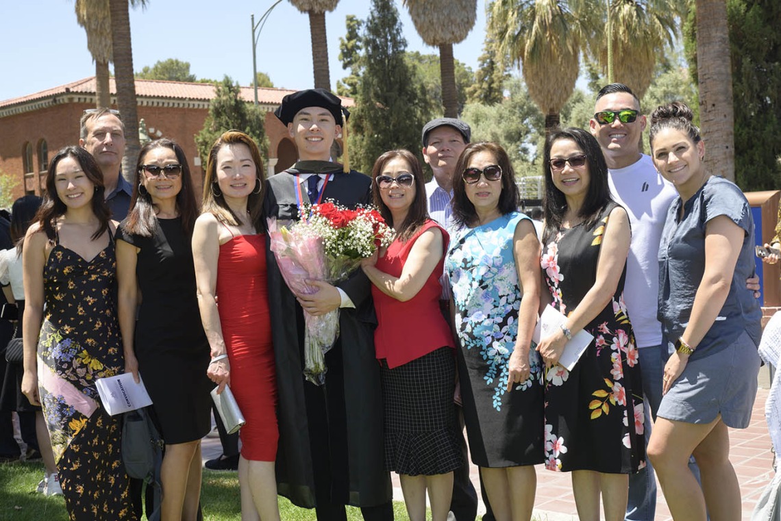 A young male graduate in cap and gown smiles while holding flowers as he stands with about 10 family members outside.