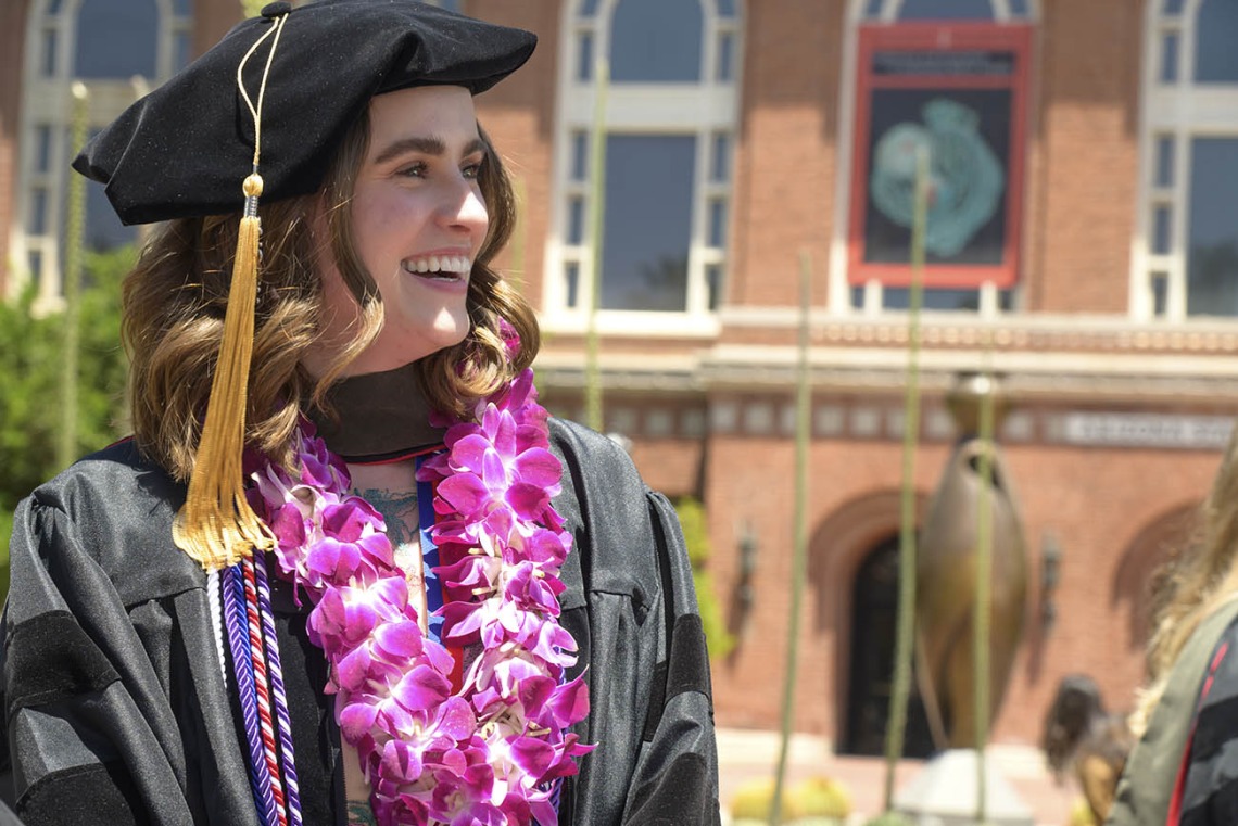 Profile of a smiling young white woman in a cap and gown with flowers around her neck with a red brick building in the background.  