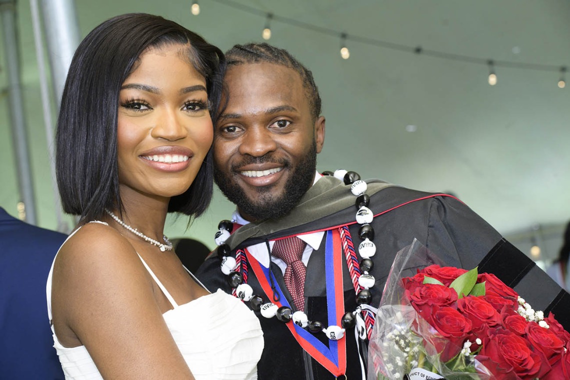 A smiling young Black man in a graduation gown holding flowers, side-hugs a young, smiling Black woman. 