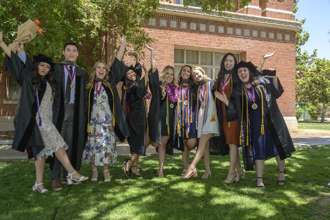 A group of nine graduates in caps and gowns stand together smiling and cheering outside. 