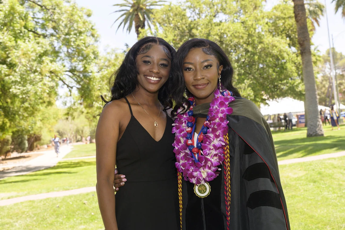 Two young Black women, one in a dress and the other in a graduation gown, side-hug and smile.