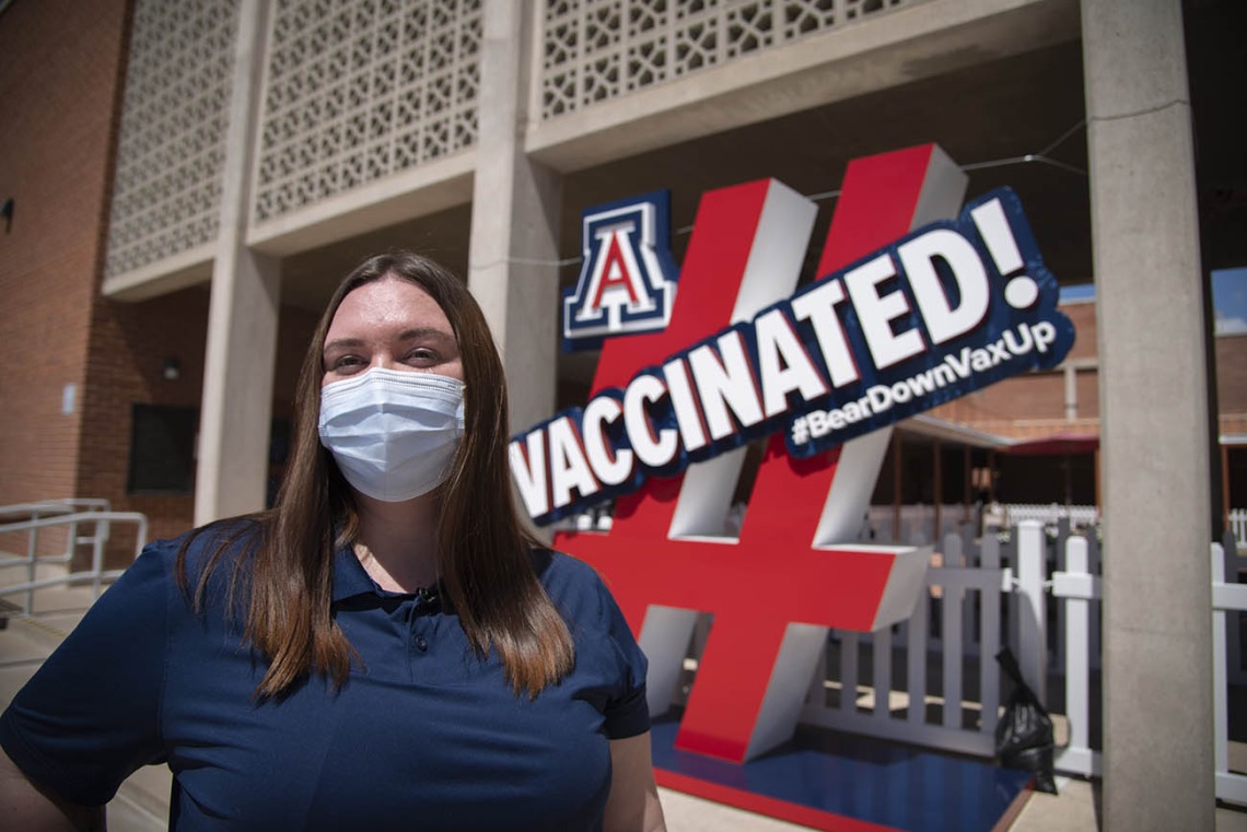 Fourth-year College of Pharmacy PharmD student Annie Hiller stands by a sign in front of the walk-in vaccination site on the University of Arizona campus. Hiller has assisted with preparing the Pfizer vaccines for distribution at the University POD.