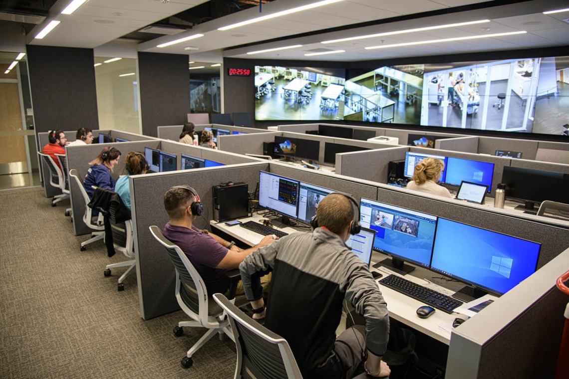 From an adjacent control room, SimDeck participants are observed on monitors.