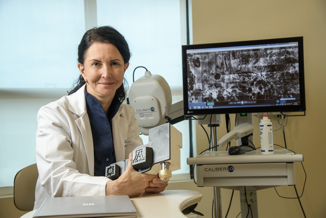 Clara Curiel Lewandrowski, MD, in front of a screen showing the images produced by a confocal microscope. She holds the smaller, portable version she is developing for wider use by doctors.